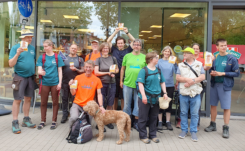 Das Bild zeigt eine Gruppe von Menschen mit einem Hund vor einem Geschäft. Sie zeigen Tüten.